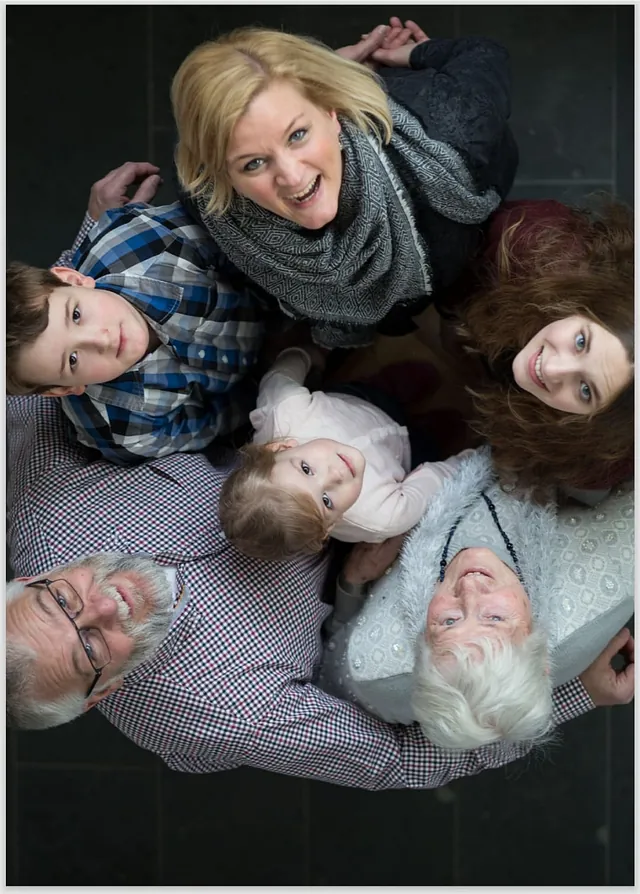Eine Gruppe von sechs Menschen unterschiedlichen Alters steht in einem engen Kreis und blickt gemeinsam lächelnd nach oben in die Kamera. Die Aufnahme aus der Vogelperspektive zeigt eine herzliche Gemeinschaft, die Verbundenheit und Lebensfreude ausstrahlt.